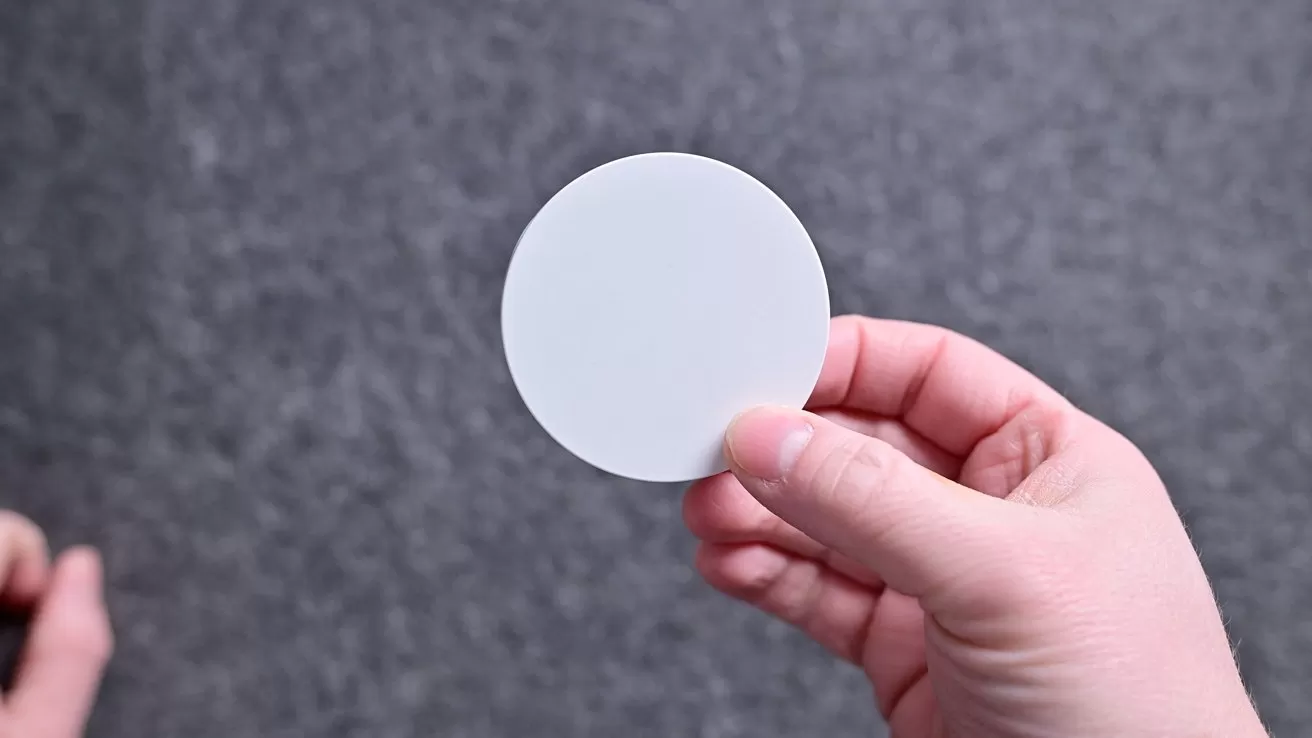 A hand holds a plain white circular disc against a gray textured background, with another hand partially visible in the lower left corner