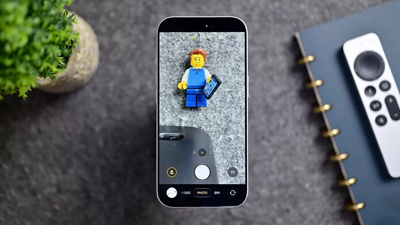 Smartphone on a gray surface photographing a small Lego-style figure, with a plant, notebook, and remote control lying nearby
