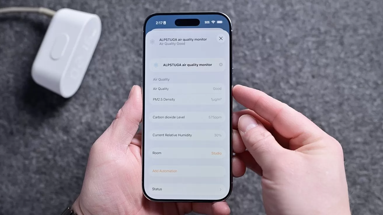 Hands holding a smartphone displaying an air quality monitor app with sensor readings, against a gray surface, with a white plugged-in device resting nearby