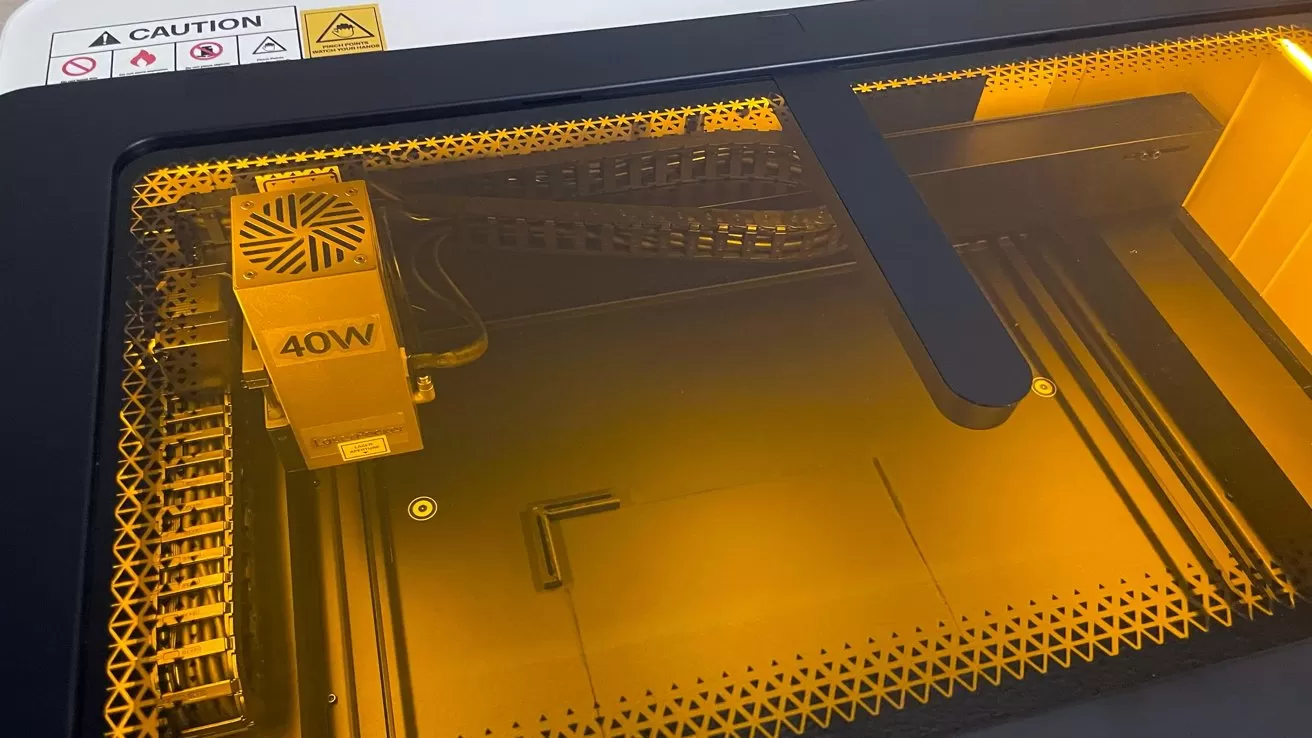 Interior of a desktop laser cutter viewed through yellow safety cover, showing a 40W laser head on rails above a patterned metal work surface with caution label on the frame