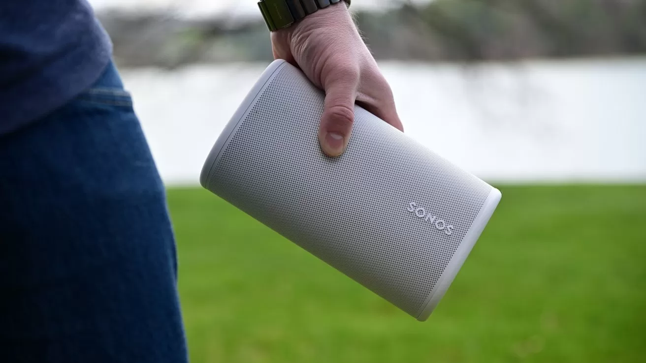 Person holding a portable white Sonos speaker outdoors near a body of water, with green grass in the background and only their hand, wristwatch, and jeans partly visible