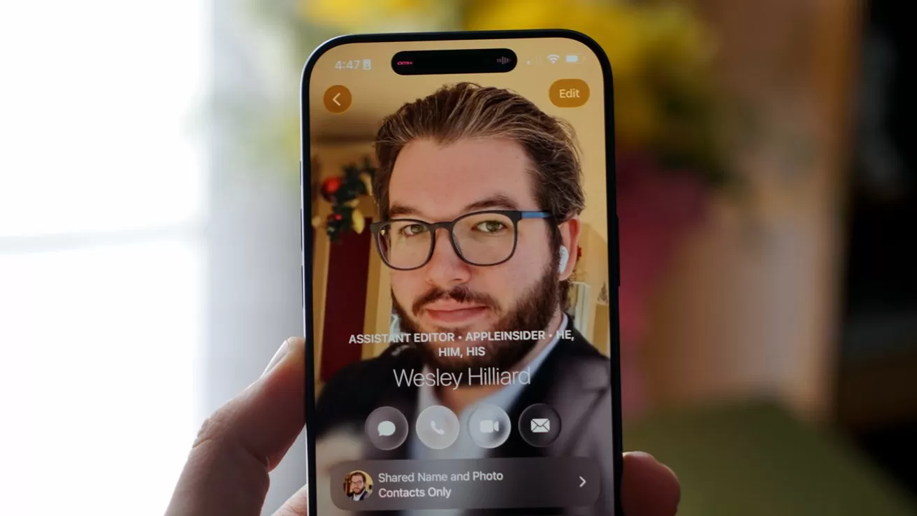 Hand holding a smartphone displaying a contact screen with a bearded man wearing glasses and earbuds, centered on the screen, with blurred indoor background and soft warm lighting