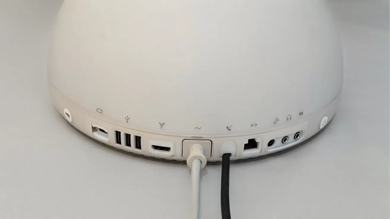 Back view of a white dome-shaped computer base showing multiple ports and cables, including USB, Ethernet, audio jacks, and power connections, resting on a light-colored surface