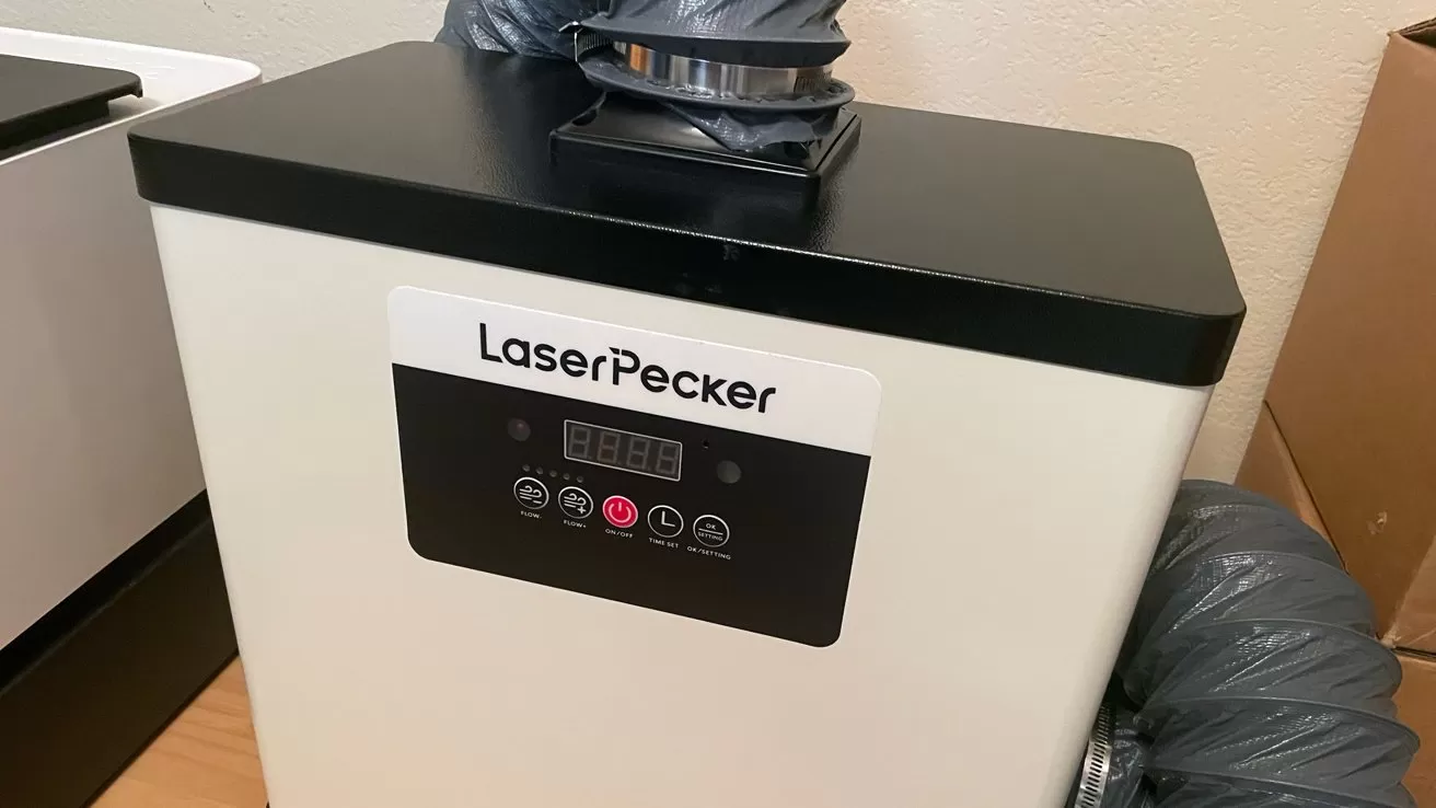 White LaserPecker machine with black top, digital control panel, and vents connected by flexible gray ducts, sitting on a wooden surface against a beige wall.
