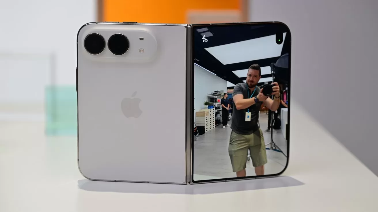 Foldable Apple smartphone partially open on a table, showing dual rear cameras on one half and a man taking a photo in the reflective screen on the other half