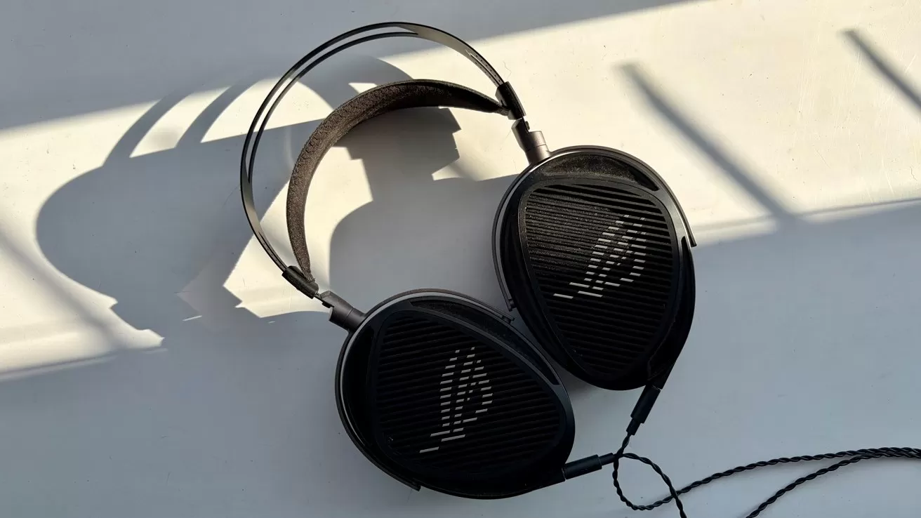 Black over-ear headphones with large oval ear cups resting on a light surface, casting long shadows from bright side lighting, with a thin audio cable extending from one side