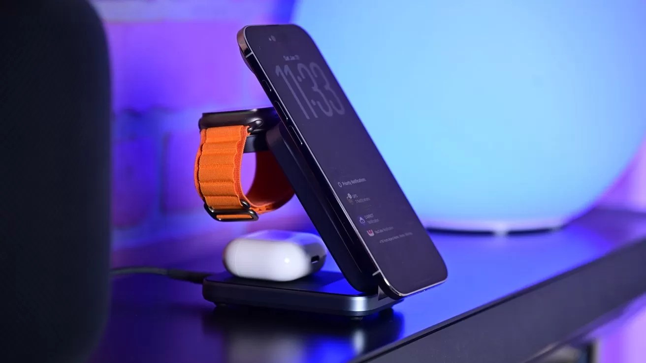 Smartphone, smartwatch with orange band, and wireless earbuds charging together on a sleek black stand, set on a desk with soft blue and purple ambient lighting