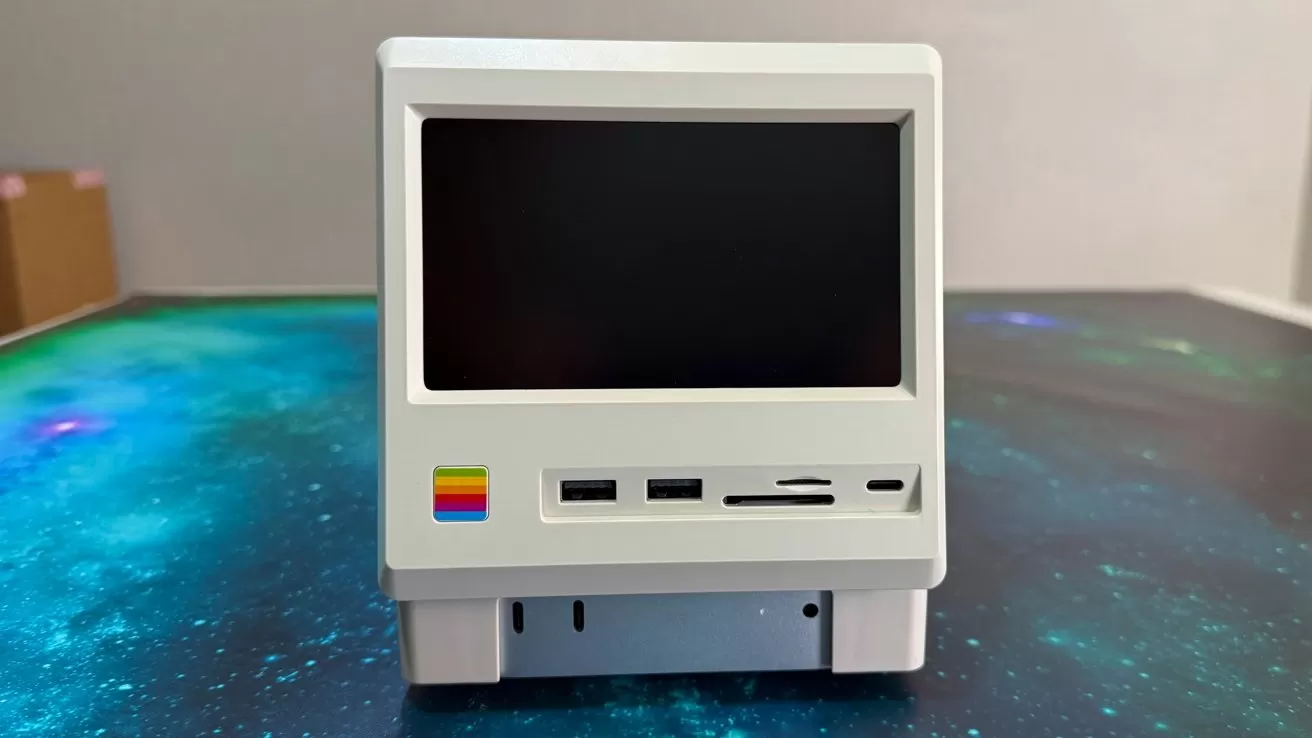 A small, white, retro-style computer with a black screen and colorful logo on a blue-green space-themed surface.