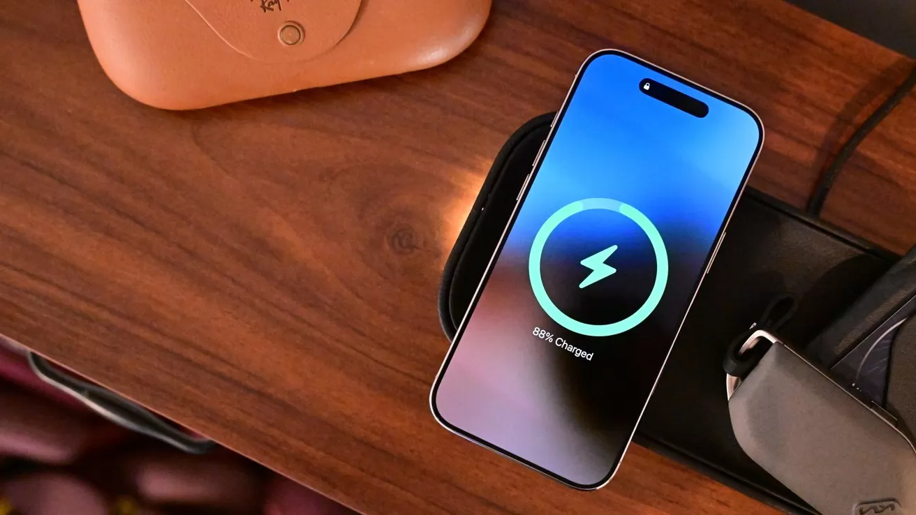 Smartphone on wooden table showing wireless charging screen with a glowing circular battery icon and percentage, next to a leather case and other small accessories
