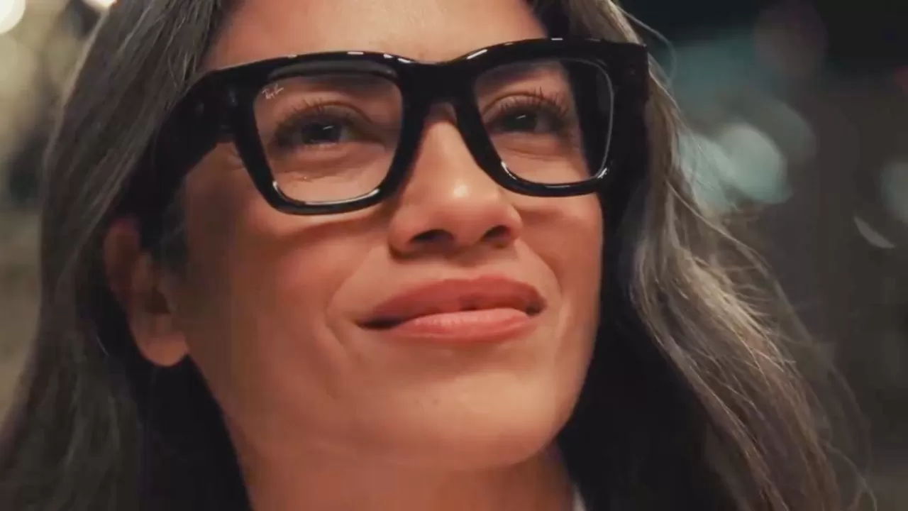 Smiling person with long dark hair and large black glasses looking slightly upward, softly lit in a close-up portrait with blurred lights in the background