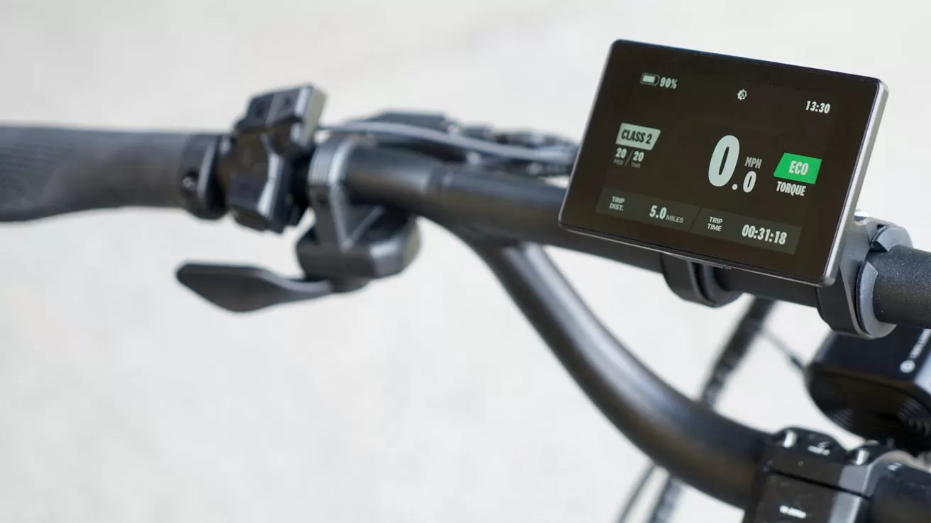Close-up of an electric bicycle handlebar with ergonomic grips, control buttons, and a digital display showing speed, battery level, eco mode, trip distance, and ride time against a blurred background