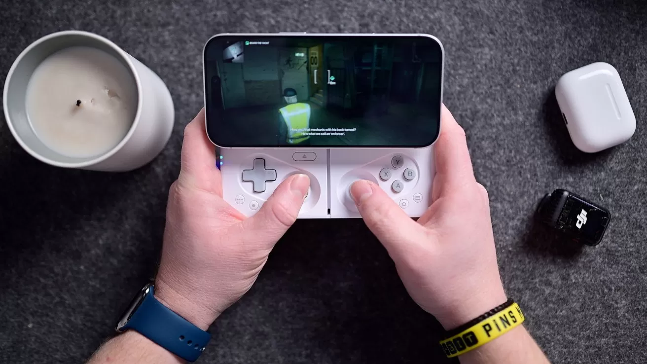 Hands holding a handheld gaming device displaying a game. Nearby are a candle, earbuds case, and a small device.