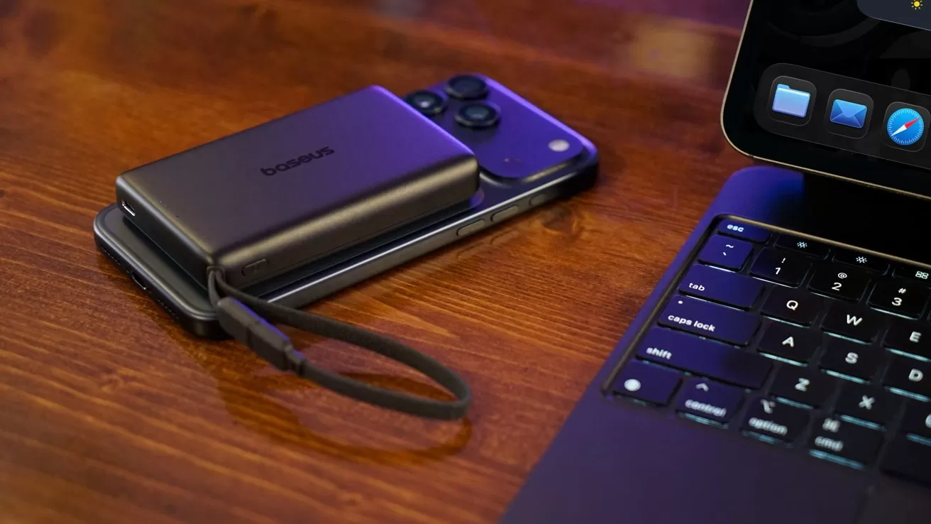 Portable power bank resting on an iPhone 17 Pro Max beside an iPad Pro keyboard on a wooden desk, lit by warm light, showing a tidy modern tech workspace