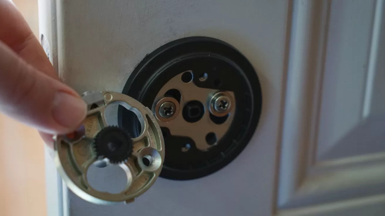 Hand holding a circular metal lock component next to a partially disassembled deadbolt mechanism mounted on a door panel