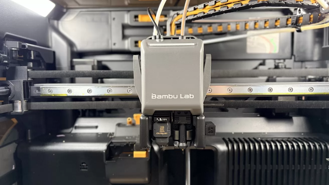 Close-up of a Bambu Lab 3D printer head inside its enclosure, showing extruder assembly, belts, rails, wiring, and surrounding mechanical components in a clean, industrial setting