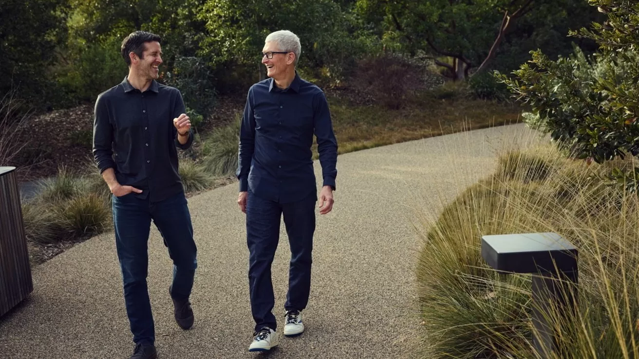Two men in casual dark clothing walk and talk along a winding gravel path surrounded by green trees and tall grasses in a landscaped park setting
