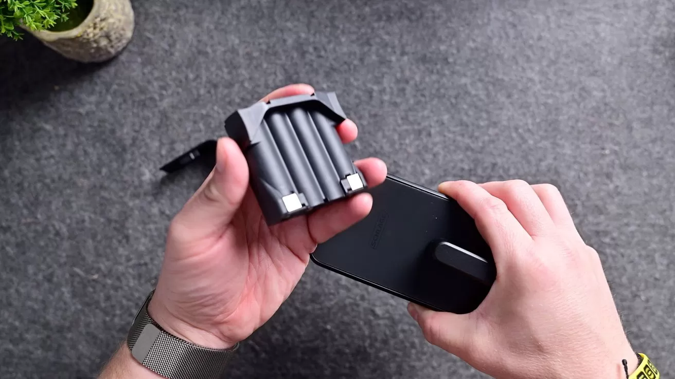 Hands holding a smartphone and a detachable battery grip with three cylindrical batteries on a dark tabletop, with a small potted plant visible in the upper left corner