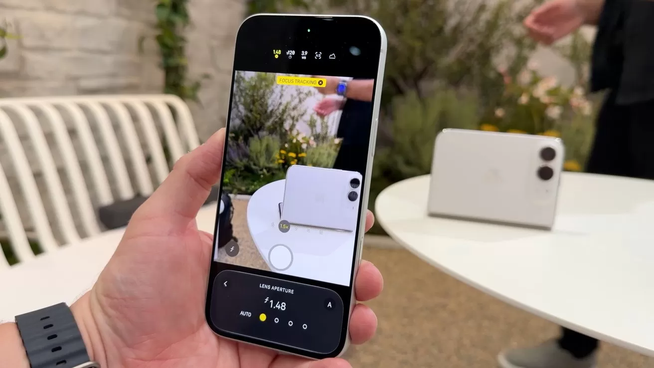 Hand holding a smartphone, camera app open, photographing a white foldable phone on a round table outdoors, with plants, a white bench, and another person's hand in the background
