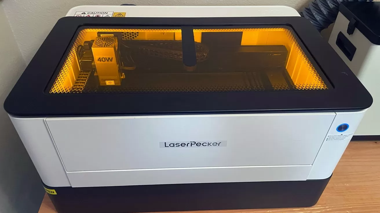 Desktop LaserPecker laser engraver with white body, dark top, and yellow-tinted viewing window, sitting on a wooden surface in a workshop or maker space