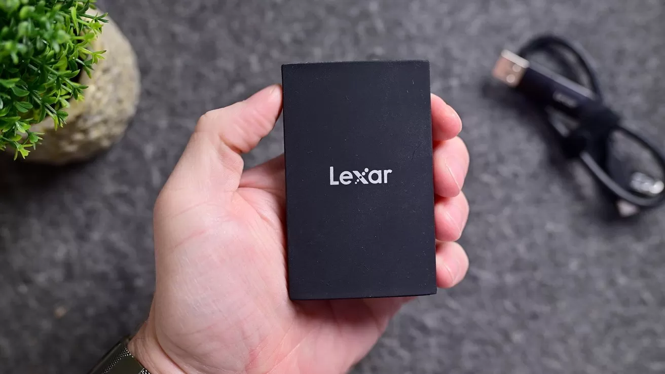 Hand holding a small black Lexar external drive over a gray surface, with a coiled USB cable and a small green potted plant in the background