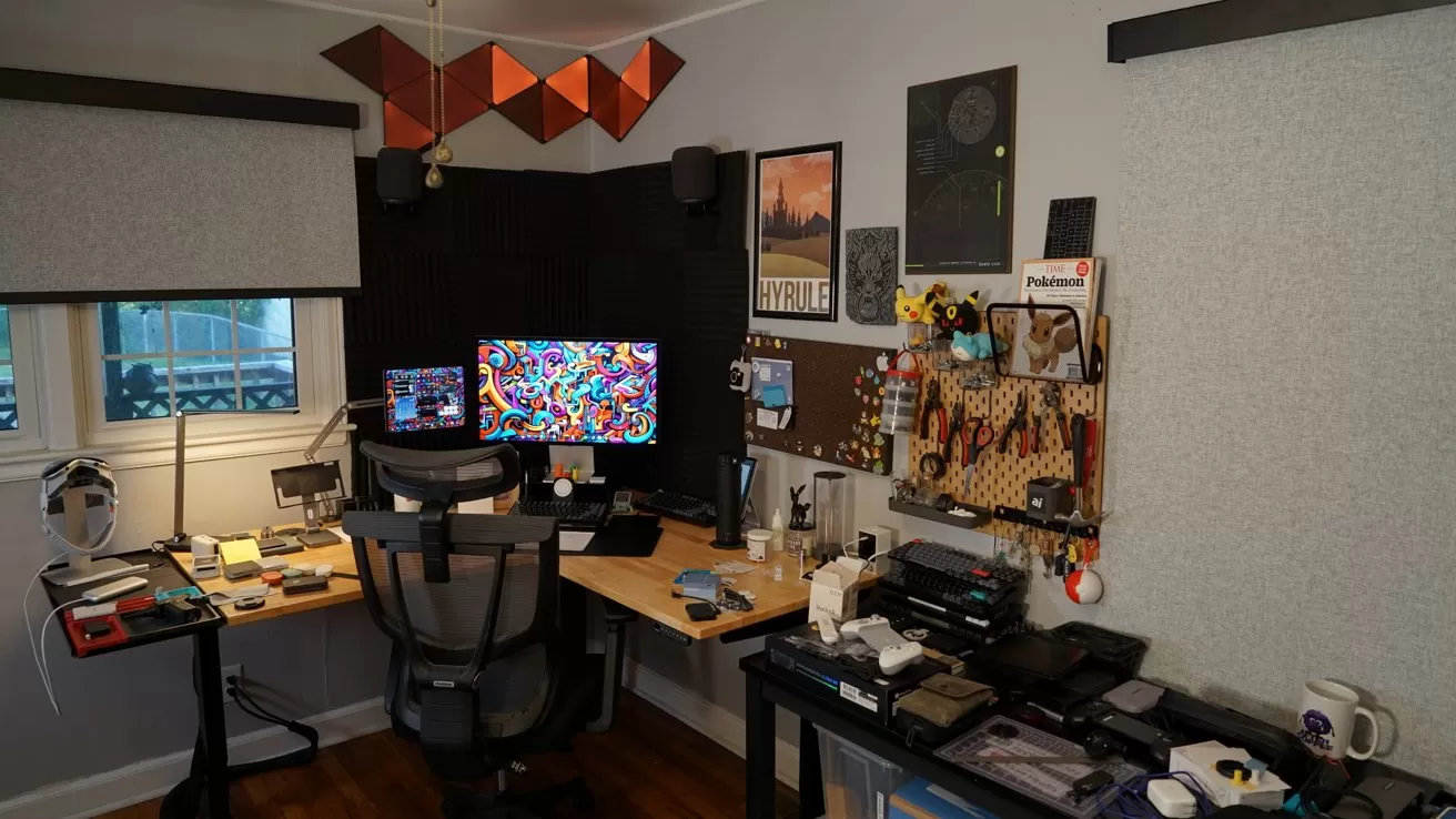 Cozy home office with dual desks, colorful monitor, ergonomic chair, pegboard of tools and trinkets, shelves of gadgets, scattered notebooks and mugs, and soft natural light from a nearby window