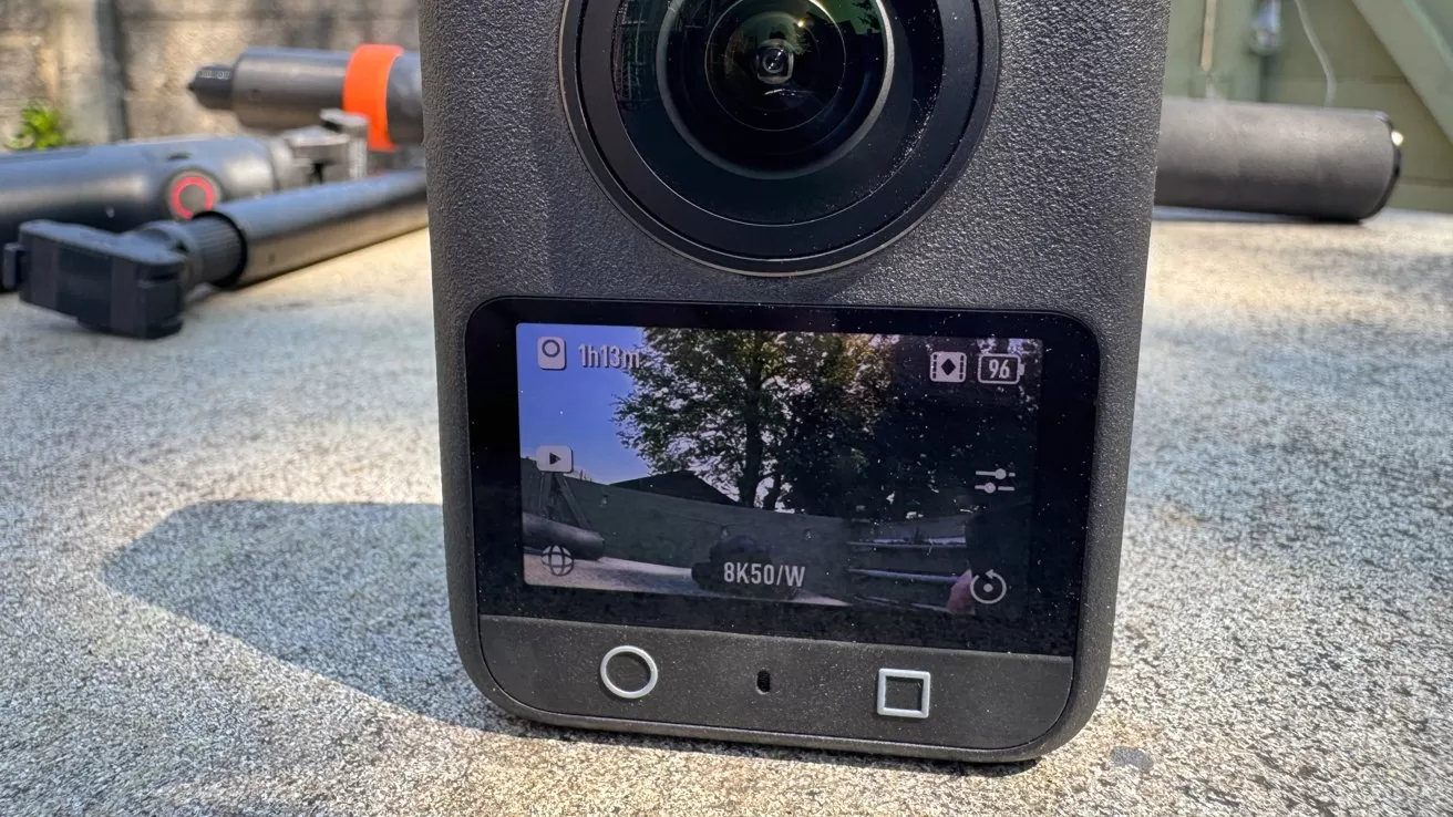 Close-up of a 360-degree action camera on concrete, showing its front lens and color screen displaying an outdoor scene with trees, battery status, recording time, and 8K50/W settings