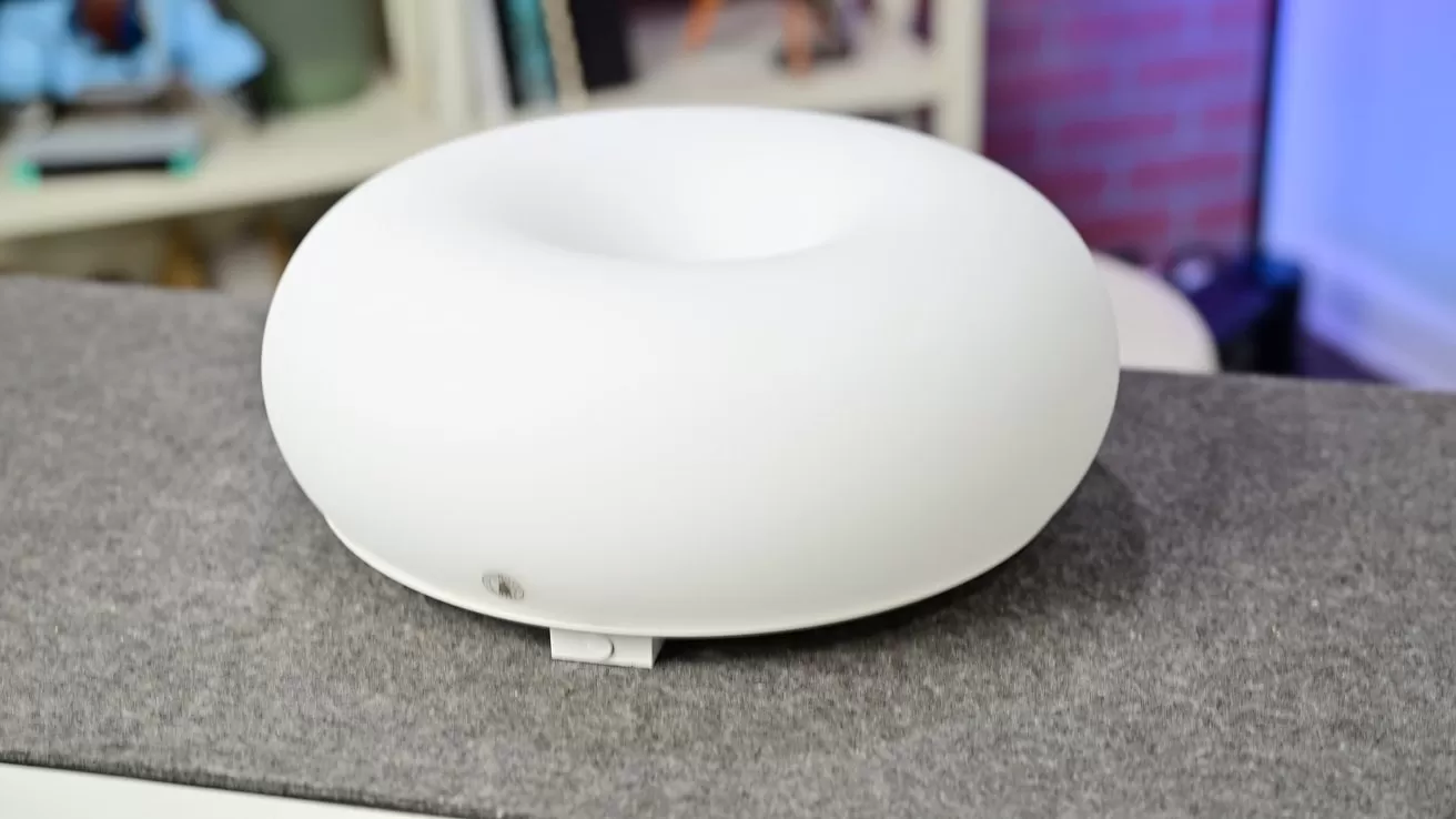 White, donut-shaped electronic device with a smooth matte surface sitting on a gray countertop, with shelves and books softly blurred in the background
