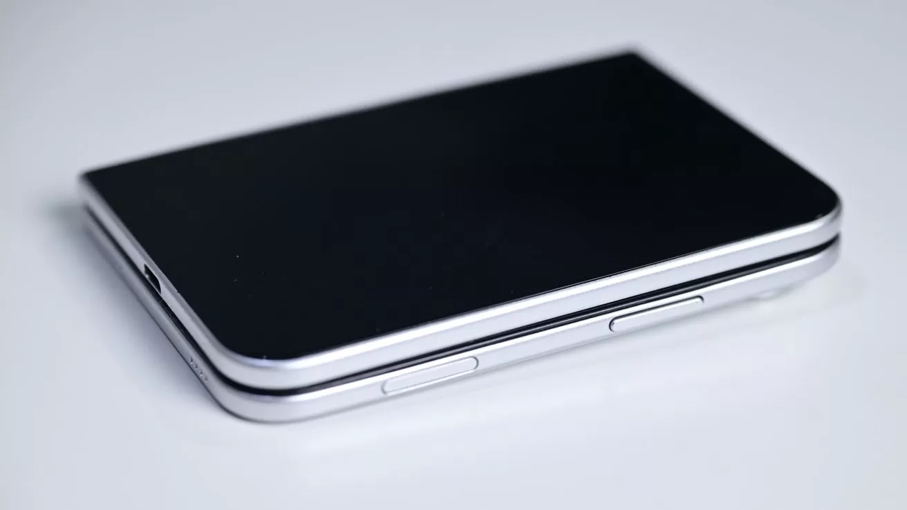 Closed silver and black foldable smartphone lying flat on a white surface, showing its side buttons and slim hinge edge