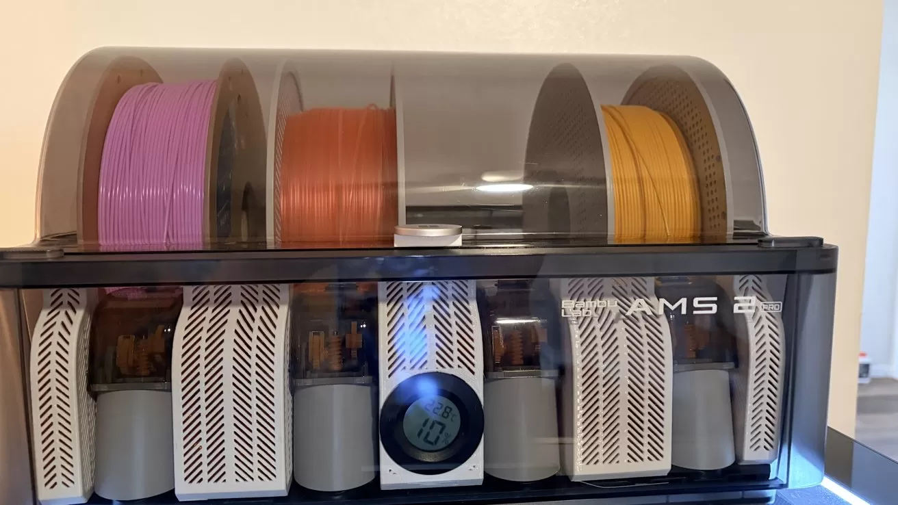 Plastic enclosure holding several colorful 3D printer filament spools, with white vented inserts and a circular digital temperature and humidity gauge on the front