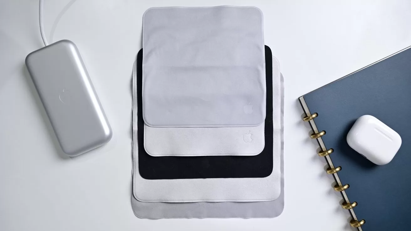 Stacked microfiber cloths in gray, black, and white beside a silver power bank, a dark notebook with gold rings, and a white wireless earbud case on a clean desk