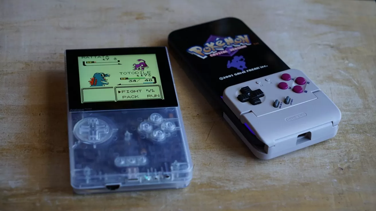 A transparent Analogue Pocket showing a Pokemon battle screen, and an iPhone in a Game Boy-style controller case displaying Pokemon Crystal title screen