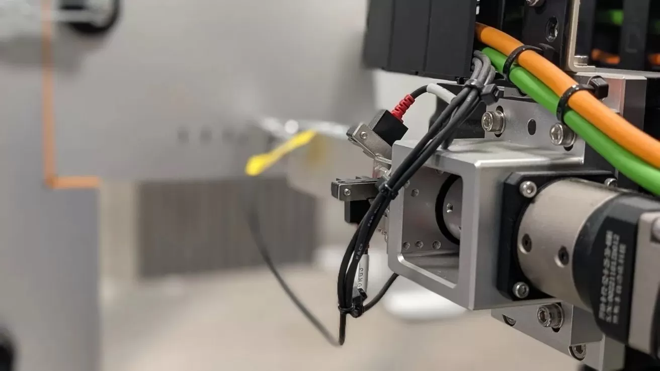 Close-up of industrial machinery showing metal components, wires, and orange and green cables connected to a motor or actuator, with a blurred gray background suggesting a factory or lab setting