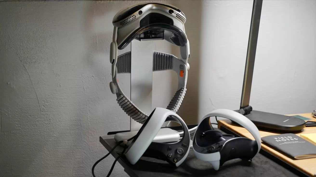Apple Vision Pro hanging on a vertical stand, with two handheld controllers resting on a desk beside a closed laptop and notebook under a desk lamp