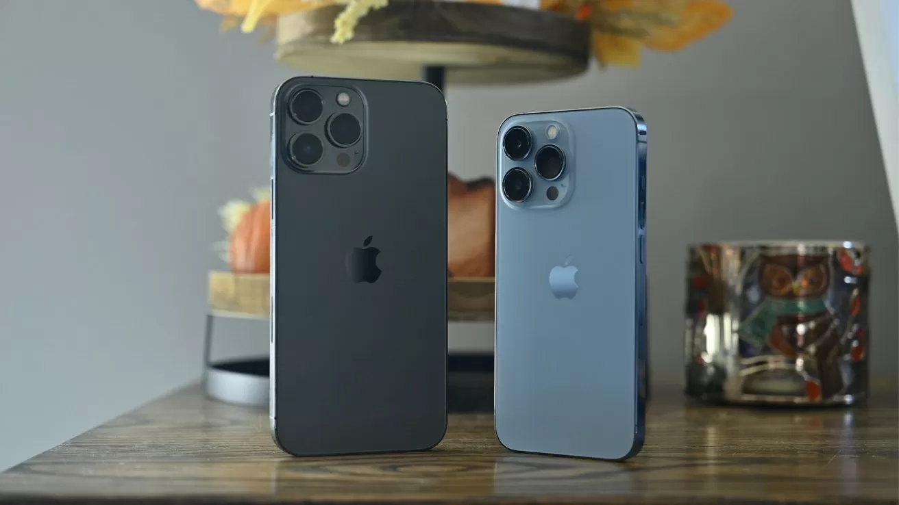 Two modern iPhones standing upright on a wooden table, one dark gray and larger, one silver and smaller, both showing triple rear cameras, with blurred home decor in the background