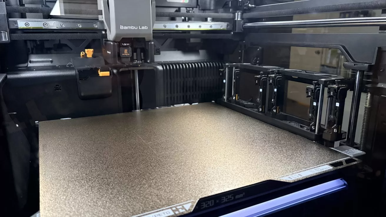 Close-up of a Bambu Lab 3D printer, showing its empty textured build plate, print head, and multiple numbered filament toolheads inside a dark enclosed printing chamber