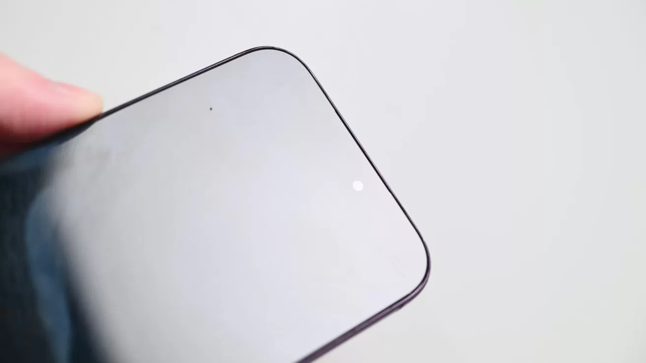 Close-up of a smartphone's top corner with the front camera and sensor barely visible on a reflective screen, held against a plain light background