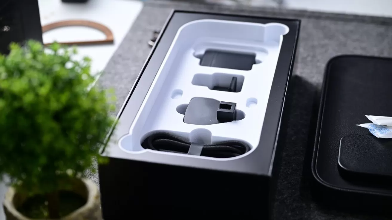 Open electronics box on a table, showing neatly arranged accessories in white packaging, including adapters and a cable, with a small green potted plant nearby and other dark items around