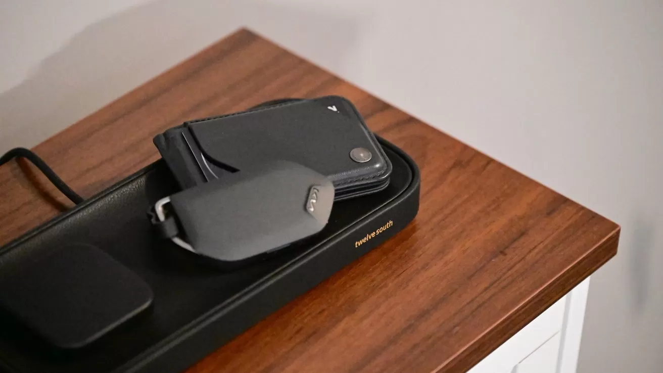 Wireless charging tray on a wooden tabletop holding a smartphone in a slim black wallet case and a compact gray electronic device with a small button