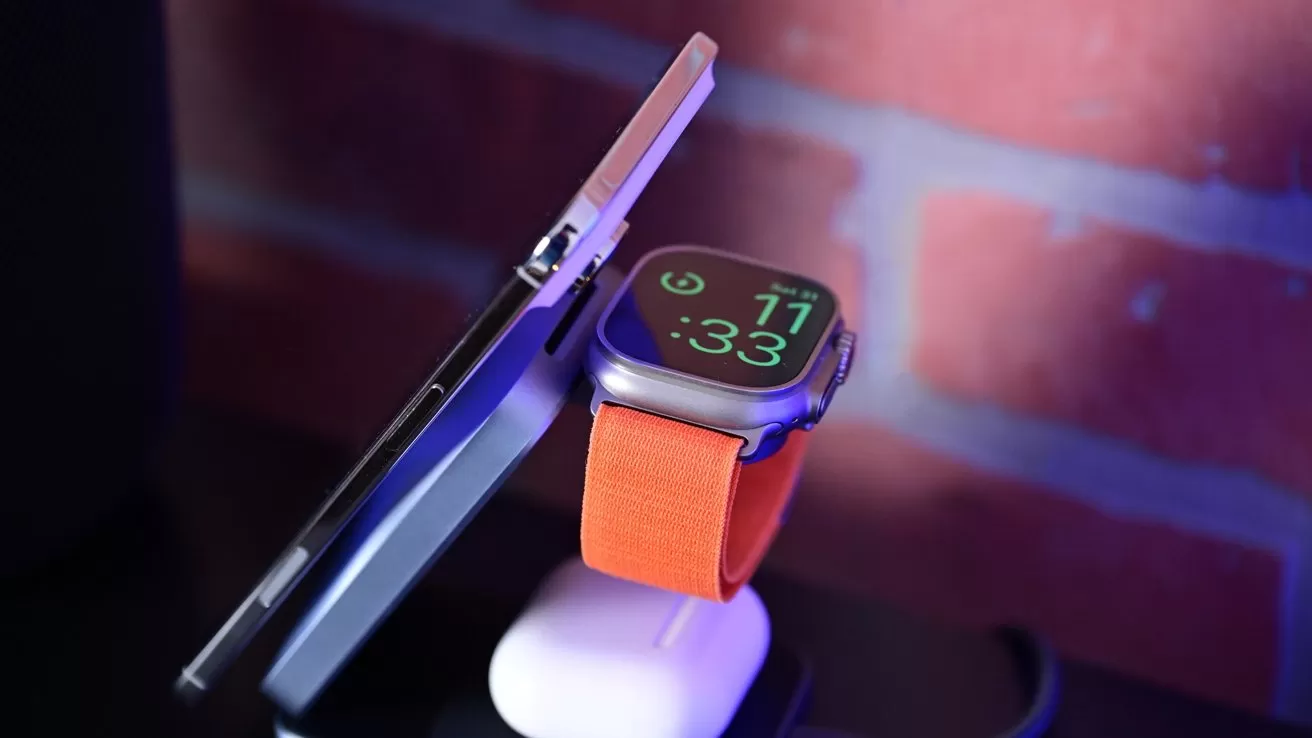 Smartwatch with orange band charging on a stand beside a vertically propped smartphone, both against a softly lit brick wall background