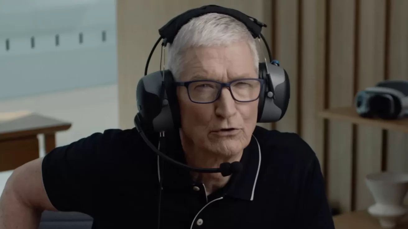 Older man with short gray hair and glasses wearing a large communication headset, seated indoors in a modern room with wooden panels and tech equipment in the background