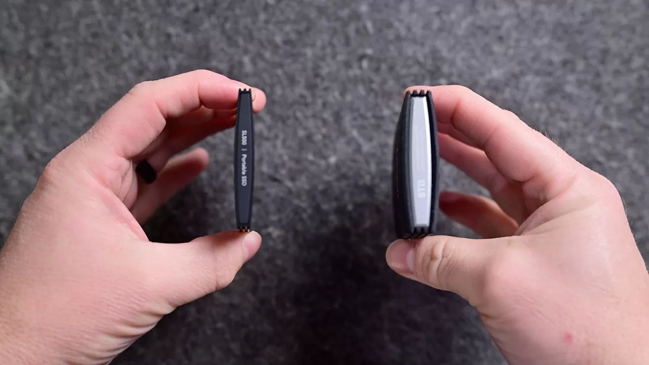 Two hands hold two small black electronic devices upright, side by side, showing a comparison between a thin flat device on the left and a thicker rounded device on the right