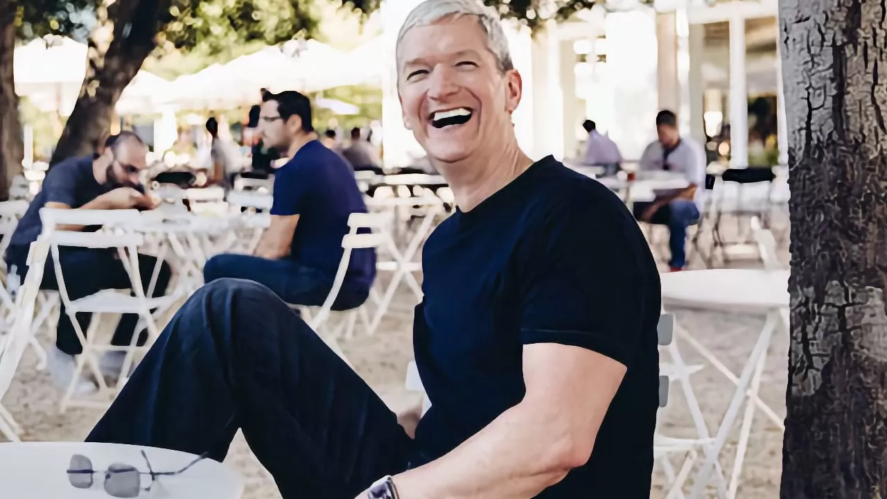Smiling man with short gray hair in a black T-shirt sits relaxed at an outdoor cafe, surrounded by people at white tables under trees on a sunny day