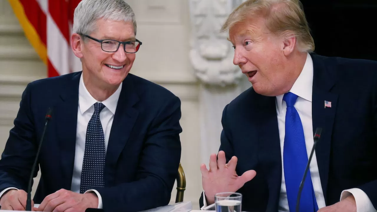 Two men in suits sit at a conference table, smiling and talking animatedly, with microphones, papers, and a glass of water in front of them and a U.S. flag behind.