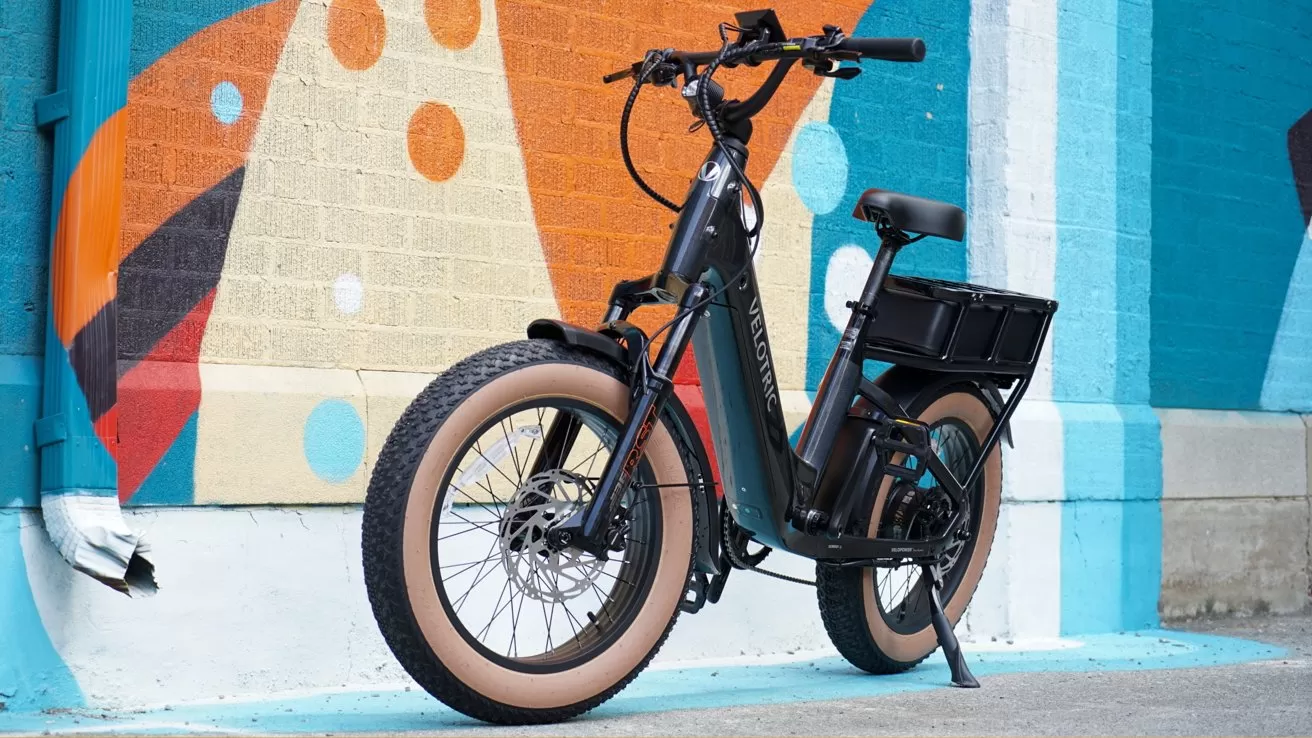 Black Velotric electric cargo bike with fat tan tires parked on a sidewalk in front of a colorful abstract mural featuring orange, blue, and white geometric shapes and polka dots
