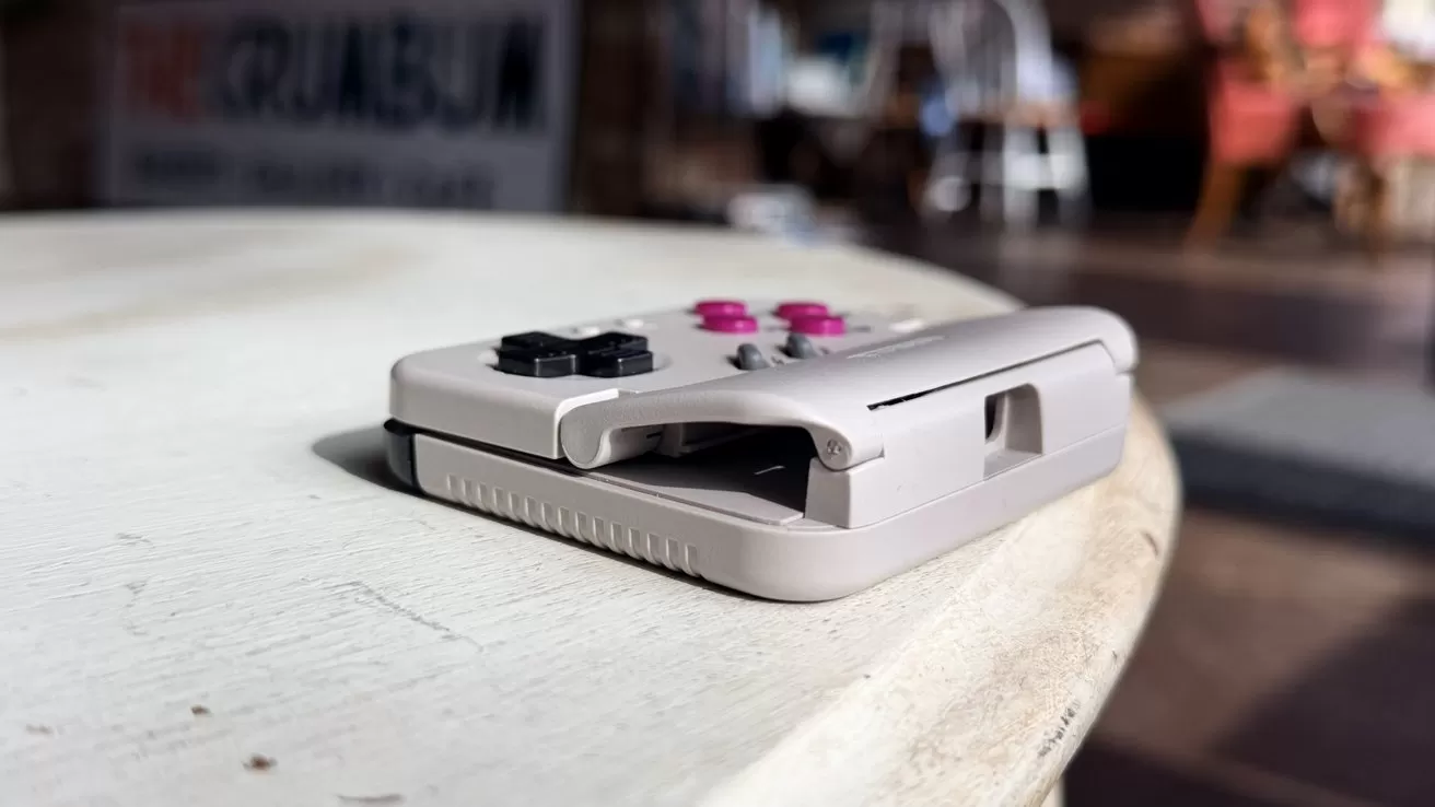 Game controller with black and pink buttons rests on a light wooden table in a softly lit cafe, background blurred with chairs and signage