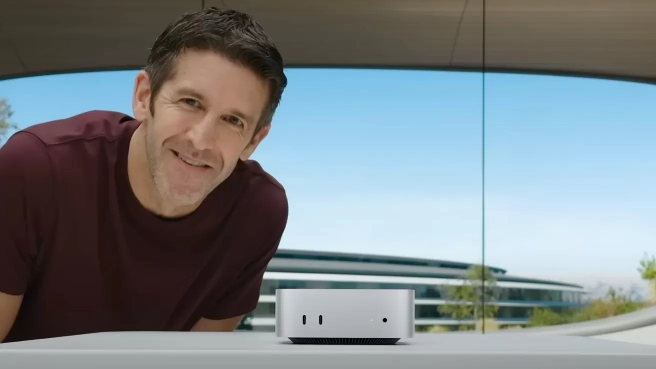Smiling man in a maroon shirt leans over a table displaying a small silver desktop computer, with a modern glass building and blue sky in the background
