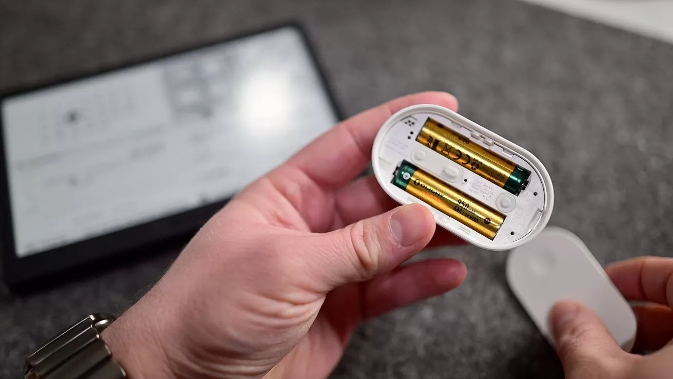 Hands holding a small oval device with its battery compartment open, revealing two AA batteries, while a blurred electronic screen or e-reader lies on a gray surface in the background