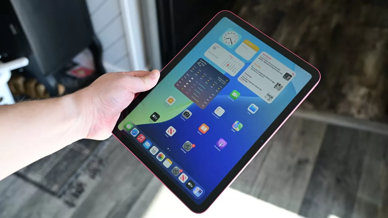 Hand holding a tablet displaying the home screen with colorful app icons and widgets, against an indoor background with wooden flooring and partial view of a doorway