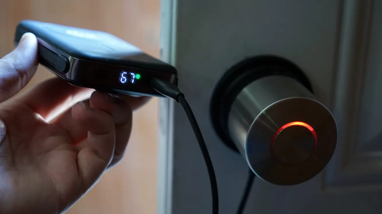 Hand holding a small digital power bank connected by cable to a modern cylindrical door lock, whose circular light glows red, suggesting the lock is charging or being powered temporarily