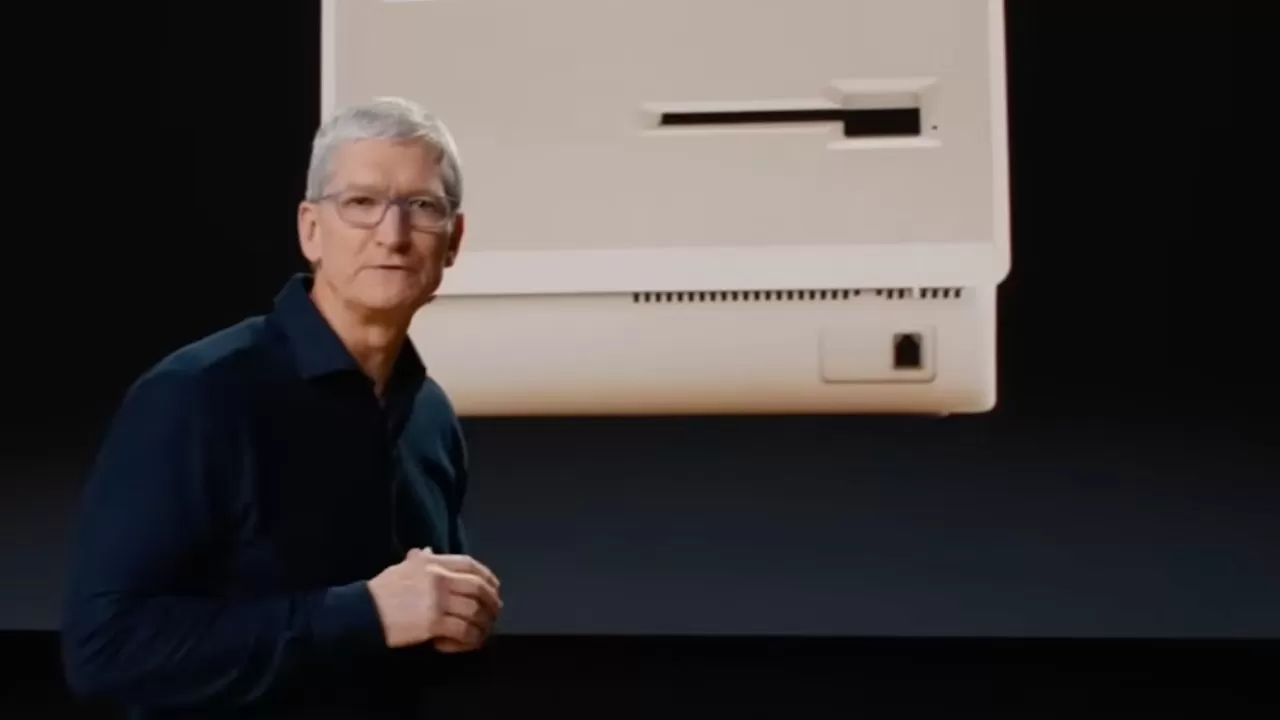 Middleaged man in glasses and dark shirt stands onstage in front of a large screen showing the beige back of a retro computer against a black background
